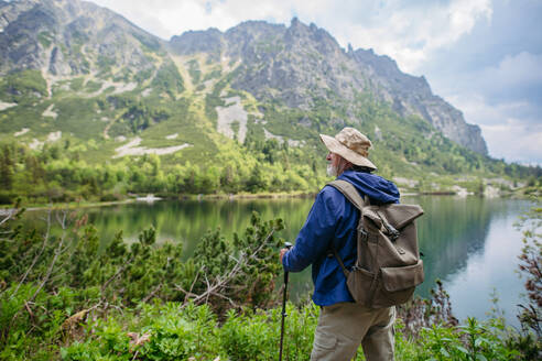 Rückansicht eines aktiven älteren Mannes beim Wandern in den herbstlichen Bergen, auf einem seniorenfreundlichen Wanderweg. Senior-Tourist mit Rucksack schaut durch ein Fernglas auf einen See. Landschaftsfotografie mit Kopierraum. - HPIF33657