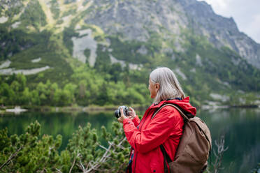 Potrait einer aktiven älteren Frau beim Wandern in den herbstlichen Bergen, auf einem seniorenfreundlichen Wanderweg. Ältere Touristin mit Rucksack, die mit einer analogen Kamera fotografiert. - HPIF33658