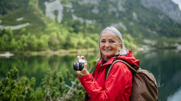 Potrait einer aktiven älteren Frau beim Wandern in den herbstlichen Bergen, auf einem seniorenfreundlichen Wanderweg. Ältere Touristin mit Rucksack, die mit einer analogen Kamera fotografiert. - HPIF33659