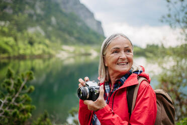 Potrait einer aktiven älteren Frau beim Wandern in den herbstlichen Bergen, auf einem seniorenfreundlichen Wanderweg. Ältere Touristin mit Rucksack, die mit einer analogen Kamera fotografiert. - HPIF33660