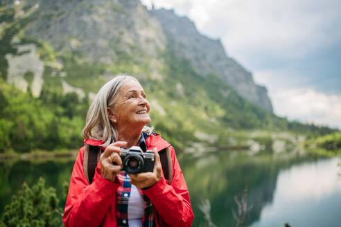 Potrait einer aktiven älteren Frau beim Wandern in den herbstlichen Bergen, auf einem seniorenfreundlichen Wanderweg. Ältere Touristin mit Rucksack, die mit einer analogen Kamera fotografiert. - HPIF33661