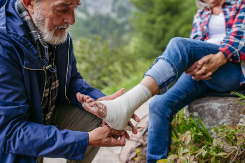 Senior man bandaging the injured leg of the female tourist. A senior woman injured her arm during hike in the mountains. Tourist went off-trail and fell. - HPIF33680