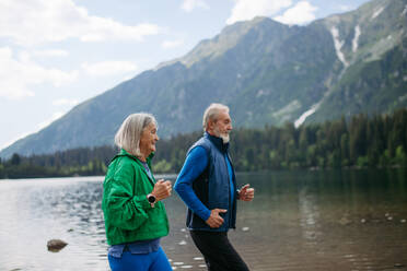 Senior couple running by the lake in the autumn. Elderly husband and wife spending active vacation in the mountains, enjoying combination of physical activity and relaxation. - HPIF33690