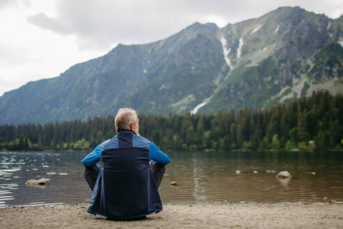 Rückansicht eines aktiven älteren Mannes, der sich nach einer Wanderung in den herbstlichen Bergen ausruht. Nachdenklicher älterer Mann, der am Ufer sitzt und die Aussicht auf die unberührte Natur genießt. - HPIF33693