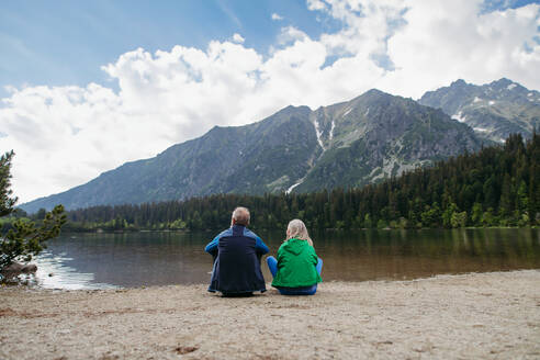 Rückansicht eines aktiven Seniorenpaares, das sich nach einer Wanderung in den herbstlichen Bergen ausruht. Nachdenklicher älterer Mann und Frau sitzen am Ufer und genießen die Aussicht auf die unberührte Natur. - HPIF33695