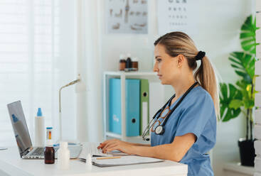 Seitenansicht einer schönen Ärztin bei der Arbeit am Laptop in einer Arztpraxis, die Papierkram und Verwaltungsaufgaben erledigt. - HPIF33705