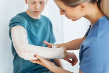 Ein Arzt prüft den orthopädischen Gips und die Klammer eines gebrochenen Arms eines jugendlichen Patienten, der sich nach einem Unfall eine Fraktur zugezogen hat. - HPIF33708