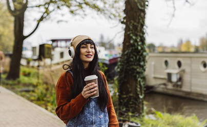 Smiling woman holding coffee cup and listening to music through wireless headphones - JCCMF10916