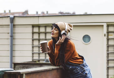 Smiling woman holding coffee cup and enjoying music through wireless headphones - JCCMF10919