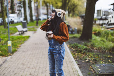 Smiling woman enjoying music through wireless headphones and holding coffee cup on footpath - JCCMF10924