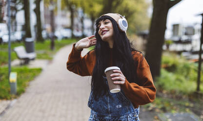 Happy beautiful woman holding coffee cup and enjoying music through wireless headphones on footpath - JCCMF10925