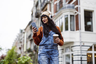 Smiling beautiful woman holding coffee cup and using smart phone in front of buildings - JCCMF10930