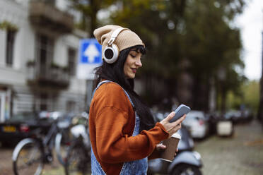 Happy woman wearing knit hat enjoying music and using smart phone on street - JCCMF10931