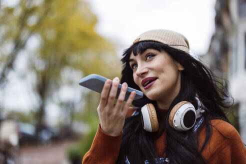 Happy beautiful woman wearing knit hat and sending voicemail through smart phone - JCCMF10934