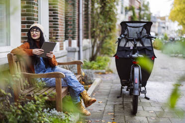 Smiling woman sitting on bench with tablet PC near cargo bike - JCCMF10940