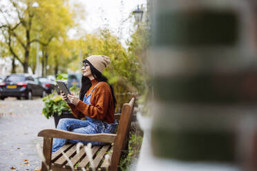 Woman wearing knit hat and sitting with tablet PC on bench - JCCMF10943