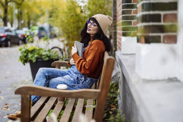 Smiling woman sitting with eyes closed and holding tablet PC on bench - JCCMF10944