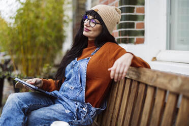 Smiling beautiful woman sitting with eyes closed and holding tablet PC on bench - JCCMF10945