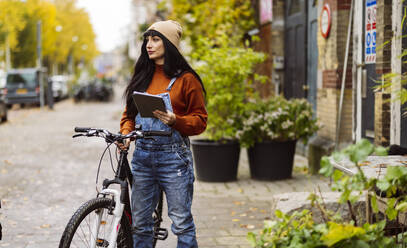 Thoughtful woman holding tablet PC and standing with bicycle on street - JCCMF10946