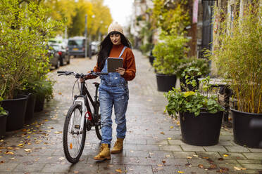 Woman using tablet PC and walking with bicycle on footpath - JCCMF10947