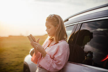 Young woman scrolling phone, leaning on her car in the nature, - HPIF33831