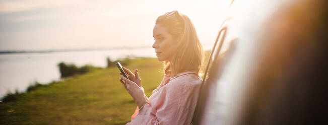 Young woman scrolling phone, leaning on her car in the nature, - HPIF33832