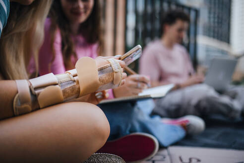 Nahaufnahme der gebrochenen Hand eines Schülers der Generation Z, der nach der Schule im Freien lernt. Der Arm trägt eine Unterarmstütze. - HPIF33900