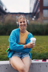 Portrait of a Generation Z girl student listening to music via wireless headphones outdoors in the city. Student spending free time alone, enjoying coffee in a reusable travel mug. Concept of gen Z as loneliest generation. - HPIF33915