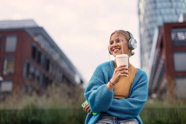 Portrait of a Generation Z girl student listening to music via wireless headphones outdoors in the city. Student spending free time alone, enjoying coffee in a reusable travel mug. Concept of gen Z as loneliest generation. - HPIF33916