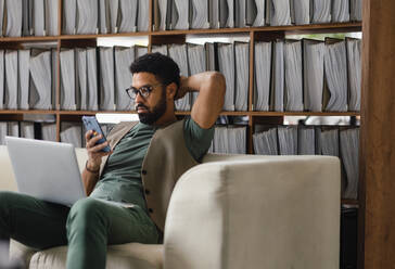 Young multiracial man sitting on a sofa and working, using notebook and smart phone in office. Manager looking at mobile phone with concerned look. - HPIF34032