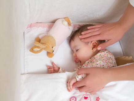 Hands of mother caressing her cute baby girl sleeping in a cot with pacifier and stuffed toy - ADSF50044