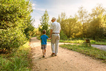 Back view of grandfather with hat and grandchild walking on a nature path - ADSF50045