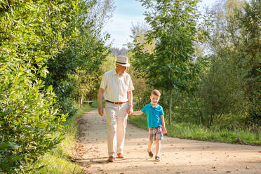 Front view of grandfather with hat and grandchild walking on a nature path - ADSF50047