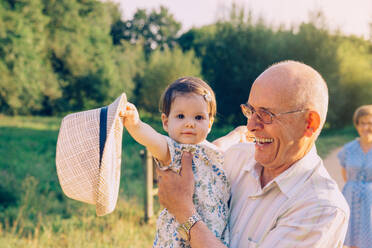 Adorable baby girl playing with the hat of senior man over a nature background. Two different generations concept. - ADSF50052