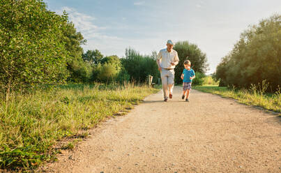 Front view of senior man with hat and happy child running on a nature path. Two different generations concept. - ADSF50054