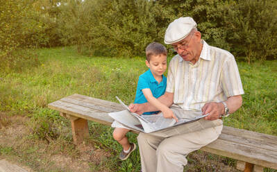 Senior man reading newspaper and cute child pointing an article with his finger sitting on park bench. Two different generations concept. - ADSF50056