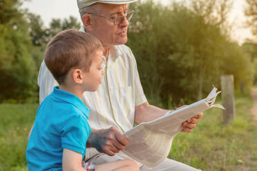 Closeup of senior man and cute child reading a newspaper sitting over a nature background. Two different generations concept. - ADSF50057