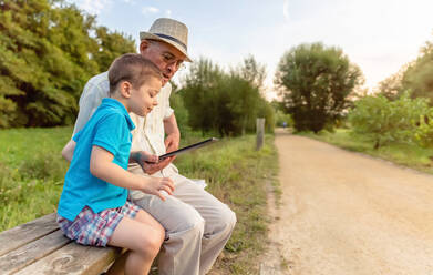 Grandchild teaching to his grandfather to use a electronic tablet on a park bench. Generation values concept. - ADSF50058