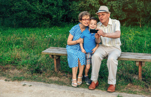 Grandfather taking selfie with grandmother and grandson sitting on a bench - ADSF50059