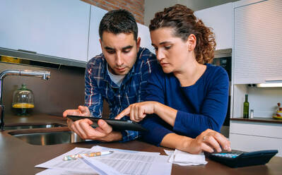 Serious young couple reviewing their bank accounts with a digital tablet and calculator at home. Financial family concept. - ADSF50064