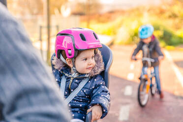 Portrait of little girl with security helmet on the head sitting in bike seat and her brother with bicycle on the background. Safe and child protection concept. - ADSF50066