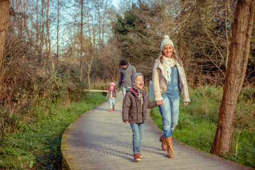 Happy family walking together holding hands over a wooden pathway into the forest - ADSF50073