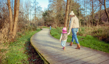 Back view of mother and daughter walking together holding hands over a wooden pathway into the forest - ADSF50074
