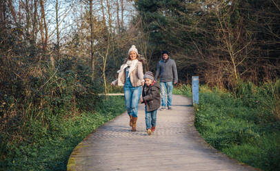 Happy kid and woman holding hands and running over a wooden pathway while man looking in the background - ADSF50075