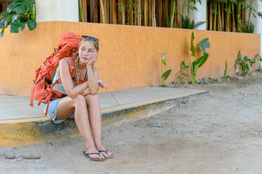 Full frame of young caucasian woman with long blond hair in casual clothes with backpack sitting on pavement and looking away thoughtfully while waiting for transportation - ADSF50083