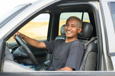 Positive young male driver in casual clothes smiling and looking away while sitting in car - ADSF50087