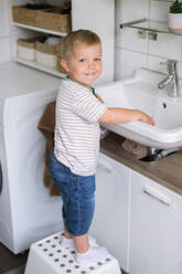 Side view of little preschool kid boy in casual clothes looking at camera while standing on stool with hands on sink near washing machine in daylight - ADSF50095