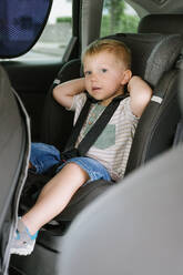 Cute preschool child boy in casual clothes looking at camera while sitting on backseat of car with open door and touching seat behind head in daylight - ADSF50101