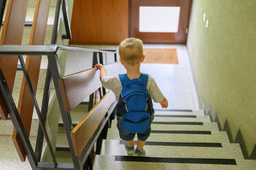 Back view of anonymous preschool boy in casual clothes with backpack looking down while climbing down steps of staircase with hand on railing at home - ADSF50102