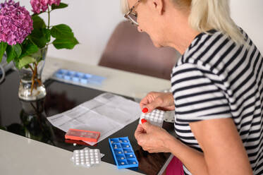 Side view of crop female in casual clothes sitting at table, sorting and putting pills and drug in weekly pill box during daytime - ADSF50105